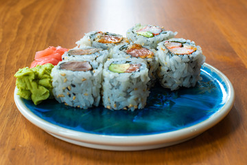 A small ceramic plate with 7 pieces of 
Japanese sushi rolls of vinegared rice, nori, avocado, salmon, tuna, ginger and wasabi. The plate sits on a wooden table. The ginger and wasabi are in clusters.
