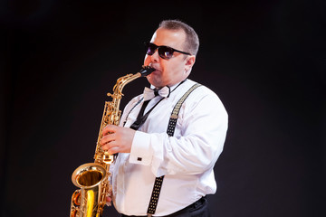 Obraz na płótnie Canvas Portrait of Expressive Caucasian Saxophonist in White Shirt Posing in Sunglasses Against Seamless Black Background.