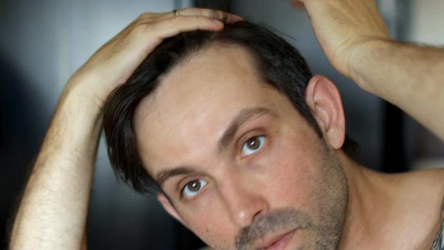 Handsome young Caucasian man looking at himself in a bathroom mirror while examining his receding hairline due to age