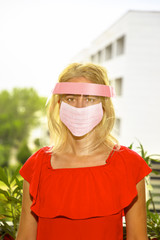 Woman with a pink surgical mask and protective visor on her face, on urban background. Concept of COVID-19 epidemic outbreak, quarantine for public healthcare and coronavirus pandemic. vertical shot
