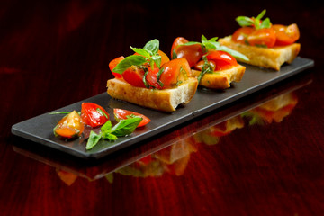 Italian tomato and basil bruschetta, classic antipasto, on baguette bread mini toasted slices. Regional mediterranean cuisine, pomodoro appetizer recipe on dark wooden background. Vegetarian food.