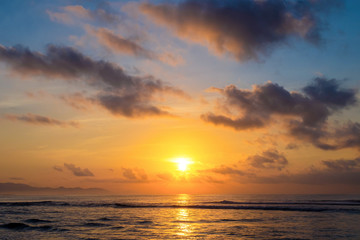 The sun rises over the ocean with backlit clouds. Beautiful sunrise at Pantai Pabean Ketewel Beach on the east coast of Bali.