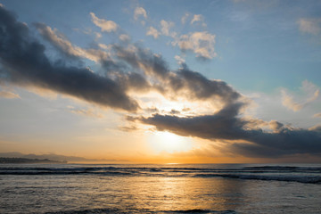 The sun rises over the ocean with backlit clouds. Beautiful sunrise at Pantai Pabean Ketewel Beach on the east coast of Bali.