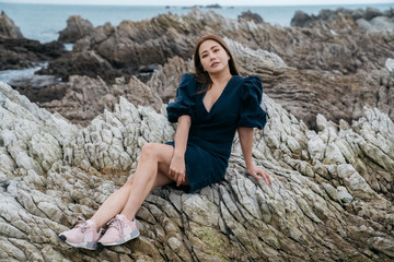 Naklejka na ściany i meble Portrait of young asian woman on stones near sea. Beautiful girl is resting on coast, enjoying outdoor recreation at Kaikoura, New zealand.