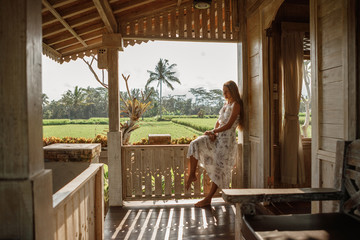 Attractive woman sitting on veranda of wooden house, relaxed summery atmosphere. Vacation on Bali, Ubud. Tropical view on background, palm tree and rice terraces