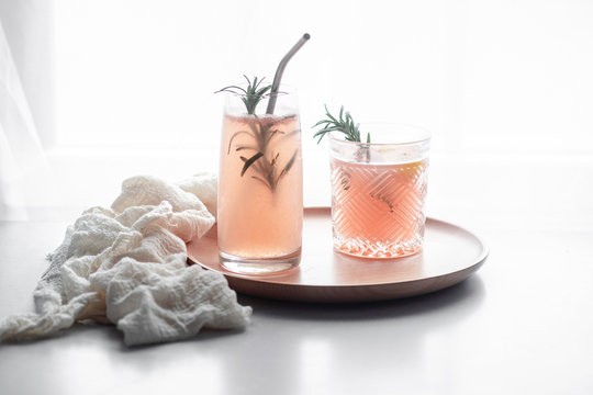 Pink grapefruit drink in two differently sized and patterned drinking glasses, garnished with rosemary and a metal straw