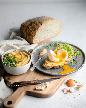 A Side Shot Of A Slice Of Bread Topped With Hummus, Herbs, And A Soft Boiled Egg, On A Silver Tray And Wood Cutting Board On A White Marble Countertop With The Cut Loaf Of Bread In The Background