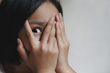 Little girl peeks through fingers, covers face with both hands, has frightened expression as...