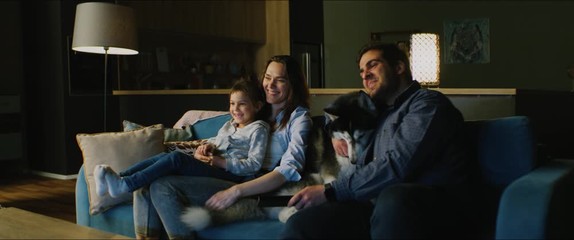 Family of three - father, mother and daughter wathcing TV in the evening. Pet husky dog lying near them. Togetherness, quarantine, stay home - Powered by Adobe