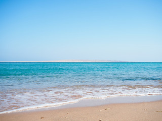Fototapeta na wymiar Red Sea photographed from beach, long view. Sea foam on brown sand. Blue sky is clear. Egypt in february, nature background. Selective soft focus. Blurred background