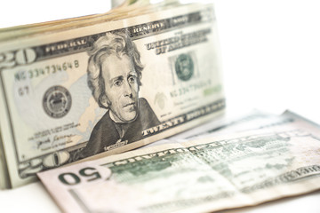 A United States twenty dollar bill stands on edge in front of a stack with a fifty dollar bill and other cash or paper currency laying on a table.