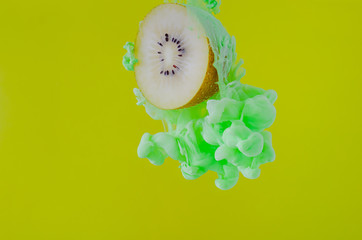 Slice kiwi fruit with partial focus of dissolving green poster color in water on yellow background for summer, abstract and background concept.