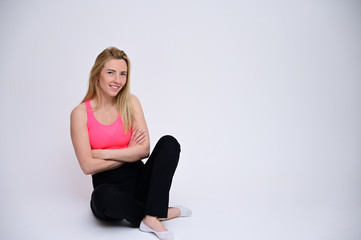 Portrait of a pretty fitness blonde girl in a sports uniform on a white background sitting on the floor. A slender Caucasian model poses with a smile.