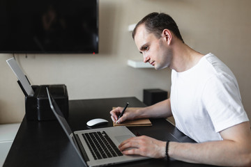 Male freelancer businessman makes notes in a craft notebook reading information in computer. Work at home during an epidemic. Self-isolation.
