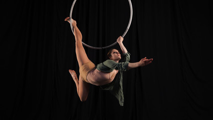 Aerial gymnast on the hoop. Young graceful girl rubber, flexibility in the air ring. Danger, risk. Female circus performer on a black background in the studio. © Artem