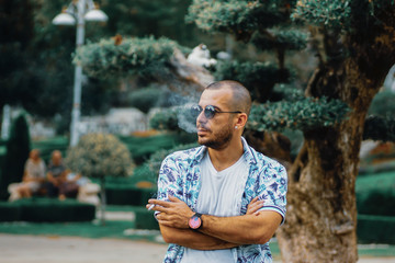 Young man smoking in the park. young man wearing a shirt with flower designs smokes cigarette outside. man wearing a white t-shirt and smoking outside. 