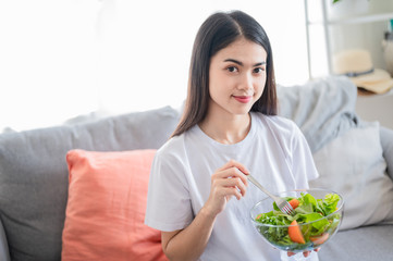 Obraz na płótnie Canvas Beautiful Asian woman are eating fresh salad she look fresh and healthy. She sit in her clear living room. New normal self care, good food for life, beauty, diet food concept.