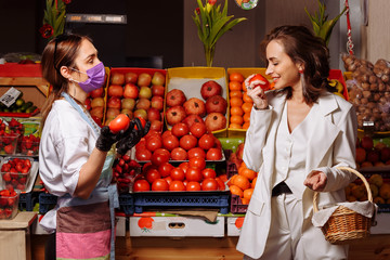 The seller in the market in a mask and rubber gloves and a young beautiful buyer girl chooses vegetables against the background of a counter with vegetables and fruits.