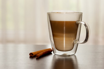 Hot coffee in clear double sided glass with cinnamon sticks on wooden table. Latte with layers