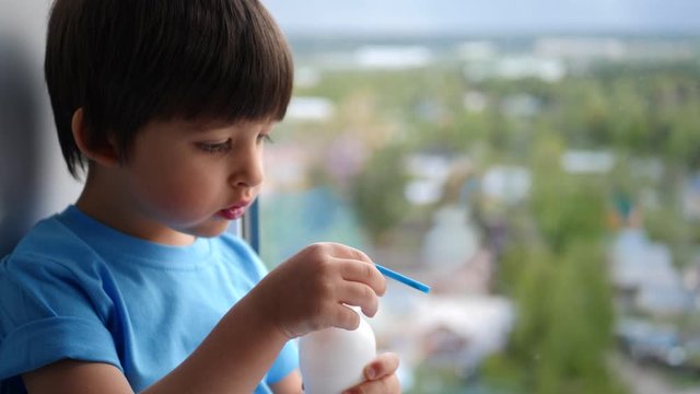 child boy in a blue t-shirt sits on the window of a high floor and drinks kefir from a plastic bottle through a straw