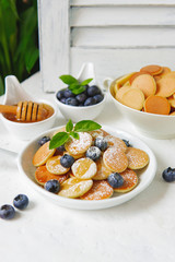 Mini pancakes with blueberries, mint, powder sugar and honey, white dishes on a white table. Beautiful breakfast, morning light. New trend dessert in social media. Little dutch poffertjes. Copy space