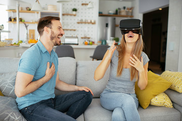 Smiling young woman using VR headset at home on couch. Woman and her husband enjoying virtual reality at her apartment.