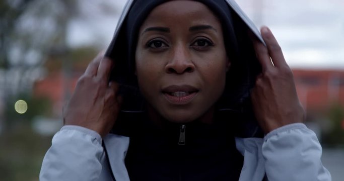 Portrait of jogger wearing running jacket puts on hood looking determined into camera and ready to run, medium close