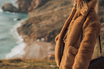 girl in a brown fur coat, in a knitted hat with a boumbon, with flying hair on a cliff, against the...