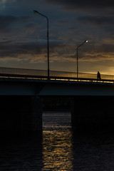 Fototapeta na wymiar Black silhouette man walked on bridge across river, yellow lantern alight his in dark dusk sunset with golden sunbeam, metafora of hope in gloomy world.