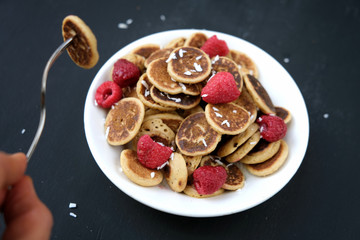 Tiny pancake cereal - trendy quarantine food. Person eating mini pancakes in bowl with fresh raspberries and coconut flakes on black background. Homemade breakfast.