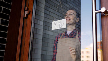 Attractive business woman owner in apron hanging, we are open sign at the front door, smiling, welcoming customers to a new cafe. Business, people and youth concept. The end of covid-19, public