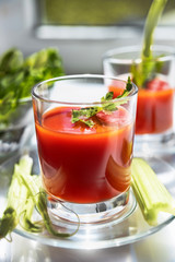 Two glasses of tomato juice with parsley and celery decorations, stand near the window, morning sunlight shines, shallow depth of field, selective focus. Natural drinks concept.
