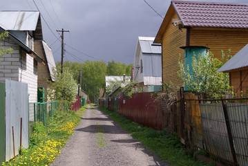 Street in a horticultural Association on a spring day. The shadow of the houses on the road. High metal fences. Grass and dandelions grow along the fence and road. On the horizon, a birch forest.