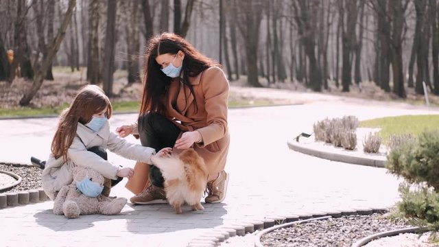 European mother and her daughter are walking with a dog. It's quarantine period so the mother and her child are wearing disposable masks to protect themselves from virus. The parent and chils are