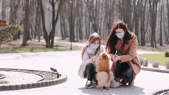 European mother and her daughter are walking with a dog. It's quarantine period so the mother and her child are wearing disposable masks to protect themselves from virus. The parent and chils are