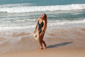 Fototapeta na wymiar Summer vacation concept. Travel caucasian woman in black bikini holdining straw hat in hands, walking on sea beach and laugh