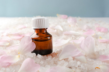 Closeup of glass bottle of essential oil, pink rose petals, white sea salt against blue background.Empty space