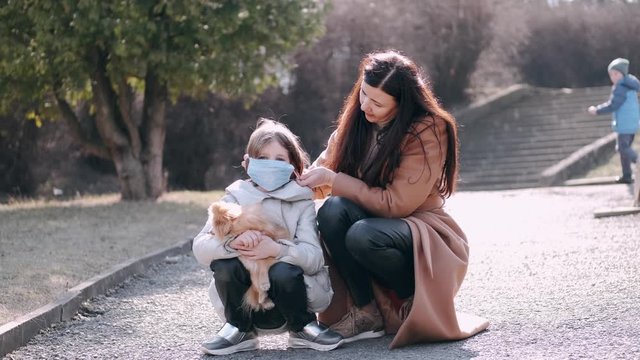 European Mother And Her Daughter Are In A Park With A Dog. It's Quarantine Period So The Mother And Her Child Are Wearing Disposable Masks To Protect Themselves From Virus. The Parent And Child Are