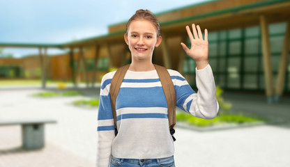education, travel and tourism concept - happy smiling teenage student girl with bag or backpack waving hand over school yard background