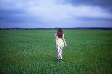 Fototapeta na wymiar Alone pretty woman walking in the field of green grass in overcast sky . Young happy girl dressed maxi dress enjoy the freedom of the meadow