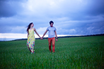 Romantic couple in relationship, walking, kissing and hugging in the countryside field of green grass in overcast sky . Young happy girl dressed maxi dress enjoy the freedom of the meadow