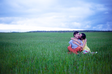 Naklejka na ściany i meble Romantic couple in relationship, sitting, kissing and hugging in the countryside field of green grass in overcast sky . Young happy girl dressed maxi dress enjoy the freedom of the meadow