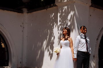 The bride and groom stand near a white wall on a light spot on the wedding day. Shadows from trees fall on faces.