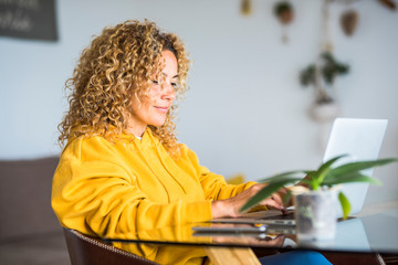 Adult pretty woman at home work with laptop computer on the table- concept of modern smart woking and job alternative office - blonde curly hair and beutiful people in technology internet connected