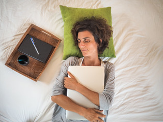 bird view of a tired woman lying in bed and hugging her laptop with eyes closed,on her stomach with pillow, tray, cup and notepad
