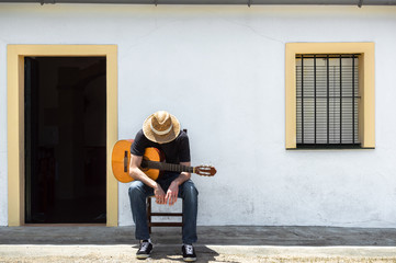 Young man sitting in front of the facade of his rural house in the countryside takes a break from playing guitar on sunny day. Rural lifestyle.