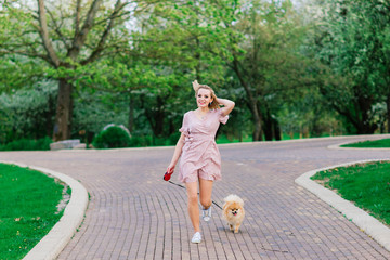 Fototapeta premium Portrait of a young glamorous female wearing pink dress holding her cute pomeranian spitz on hands. . Concept friendship between dogs and humans.