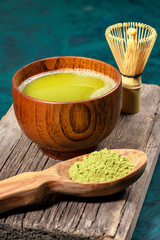 Green matcha tea in wooden cup, spoon with powder and whisk on old wooden board on emerald background. Vertical photo.