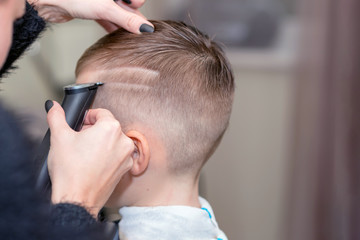 Fototapeta premium Hairdresser makes a stylish hairstyle. The woman is standing and making haircut for blonde boy. Close up of hands.