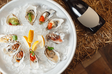 many different oysters lie on ice, a bottle of champagne on a background of hay